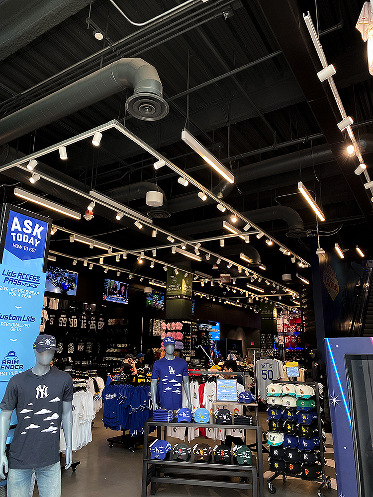 Suspended Total Track Lighting modern track system illuminating baseball caps, t-shirts, and sports merchandise displays in a Lids retail store